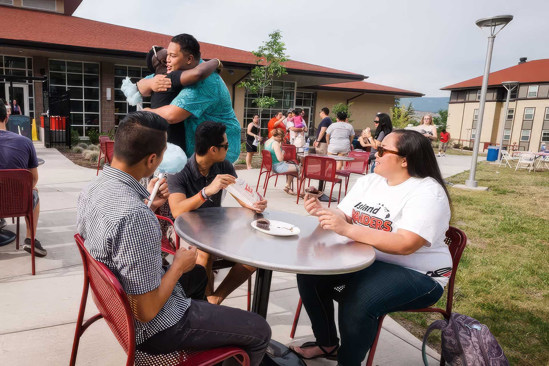 Students sitting outside at tables, eating, conversing and two individuals hugging in background